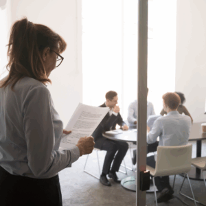 Young woman in business casual standing outside a glass-walled meeting room, holding a notebook and looking uncertain, slightly isolated, while colleagues sit inside the room